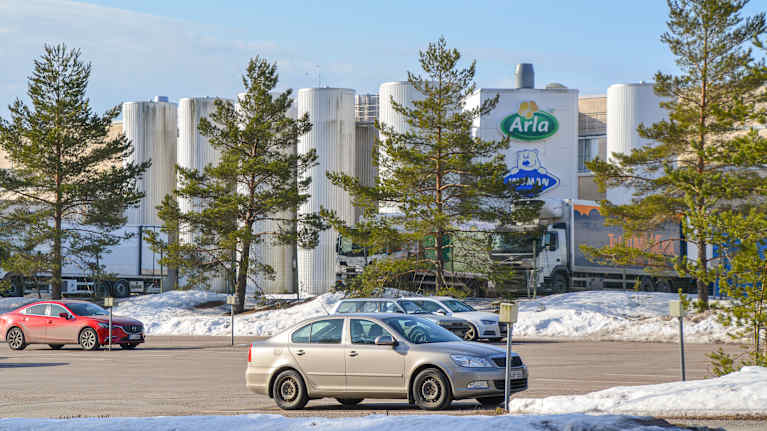 En parkeringsplats med en fabriksbyggnad i bakgrunden som har logotyperna för Arla och Ingman på väggen. 