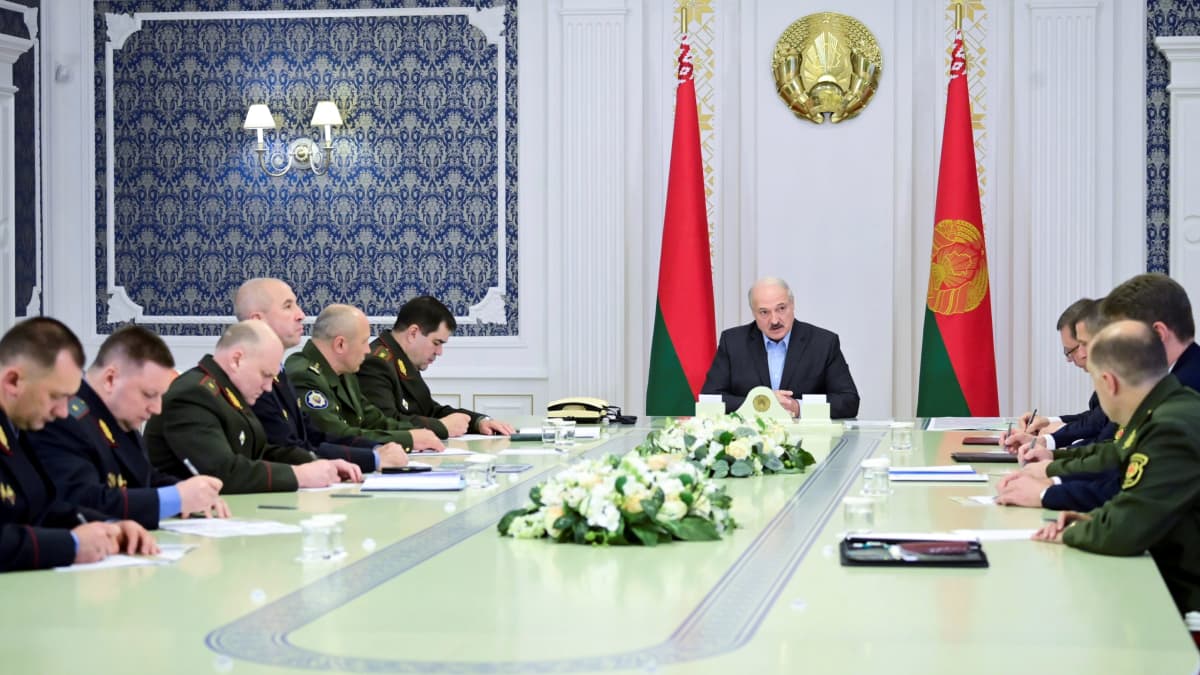 Lukashenka istuu pitkän pöydän päässä tummassa puvussa ilman kravattia. Pöydän sivuilla istuu miehiä univormuissa.