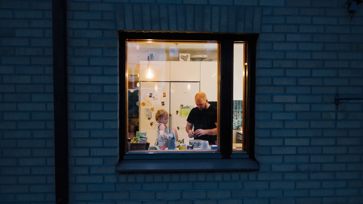 Jarkko Mandelin kokkaa keittiössä tyttärensä kanssa kuvattuna ikkunan läpi talon pihamaalta.