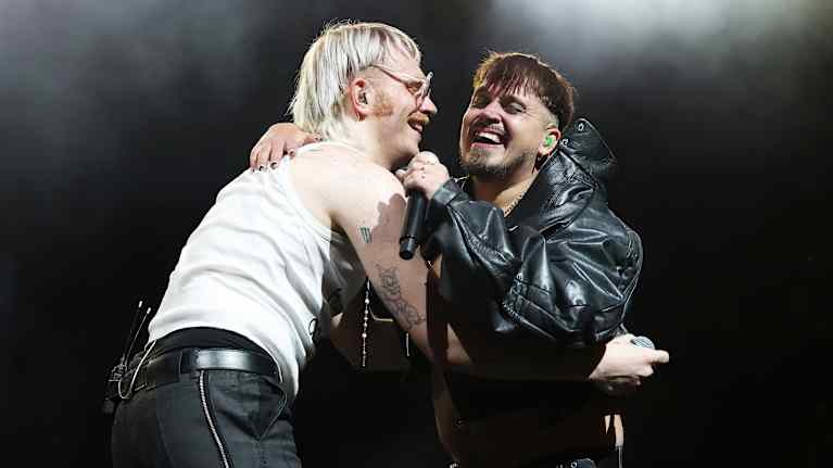 Two men onstage, one in a white sleeveless t-shirt and the other in a black leather jacket, holding microphones while embracing and smiling.