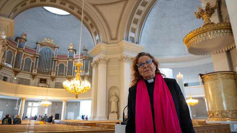 Vanhempi nainen papin asussa seisoo kirkossa. Taustalla näkyy koristeellisia kaariholveja ja näyttävät urut parvella.