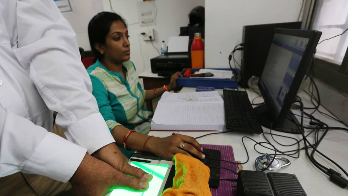 Intialainen mies laittaa peukalonsa digitaaliseen lukulaitteeseen. Intialainen nainen seuraa prosessia tietokoneelta. 
