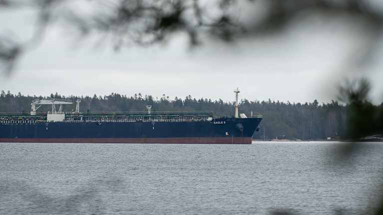 A long, low dark blue and dark reddish ship docked with a forested shore behind it.