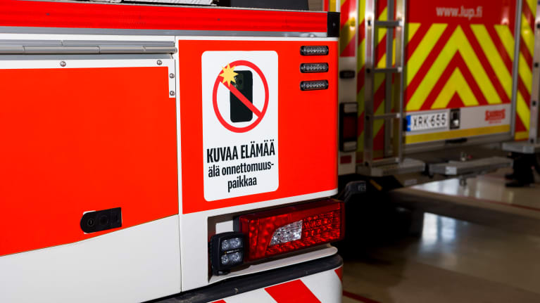 Paloauton perä, johon on kiinnitetty tarra, missä pyydetään &quot;kuvaa elämää, älä onnettomuuspaikkaa.&quot; Taustalla on toinen pelastusauto, jossa kirkkaanvärinen kuviointi. 