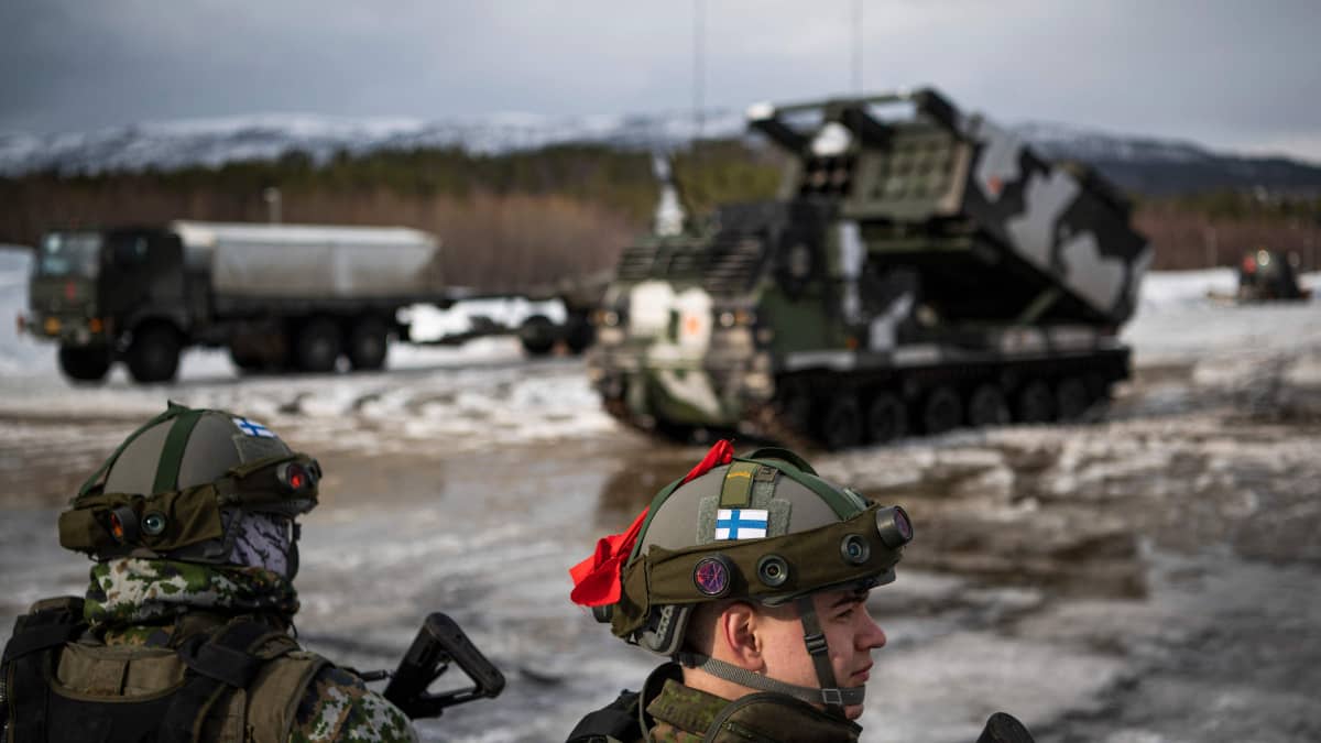 Sotilaan kypärässä näkyy Suomen lippu, taustalla raketinheitin ja muita sotilasajoneuvoja.