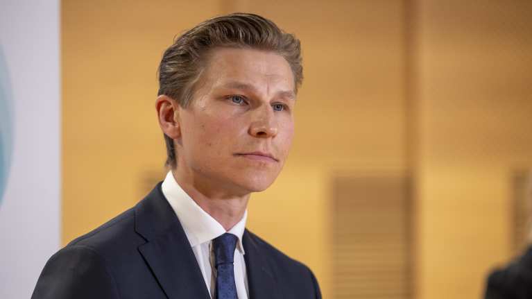 A man with a blue suit and tie and brown hair peering to the right in semi-profile.