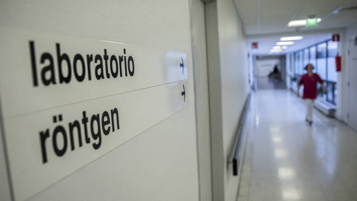 Tunnistamaton sairaanhoitaja käveleen sairaalan käytävällä. Etualalla tarkennettuna laboratorio ja röntgen kyltit seinässä.
