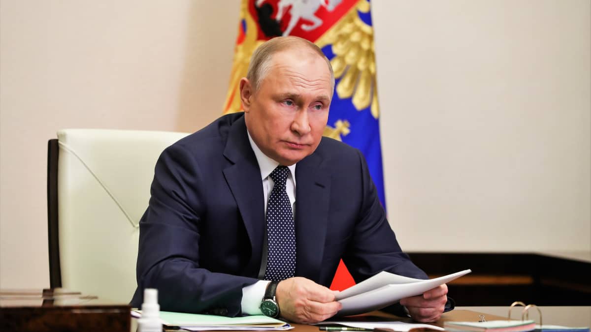 Vladimir Putin istuu vakavana tummassa puvussa ja pilkullisessa kravatissa pöydän ääressä pidellen papereita käsissään.