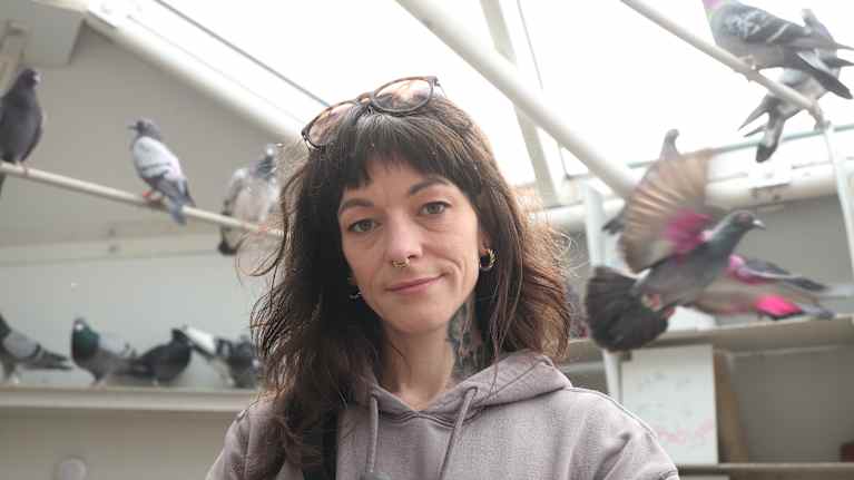 Tumma pitkähiuksinen nainen katsoo harmaata lintua sylissään pitäen kameraan. Takana näkyy lintuja orsilla ja yksi lentää siivet levällään ohi.