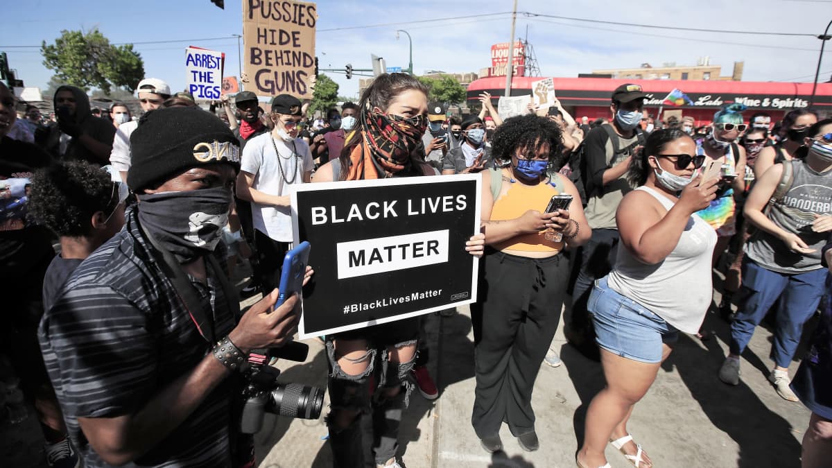 Joukko mielenosoittajia seisoo kadulla. Kuvan keskellä nuorella naisella on kädessään kyltti, jossa lukee "Black lives matter". Monilla on käsissään kännykät, joilla he kuvaavat. Monilla on myös kasvomaskit kasvoillaan.