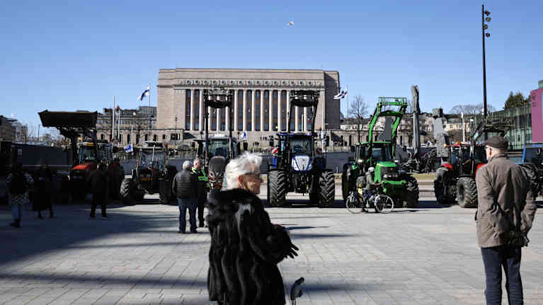 Nainen turkissa katselee eduskuntatalon edustalle kerääntyneitä traktoreita.