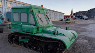 Vanha vihreä lumitelaketjullinen pieni ajoneuvo. Takana ‌puinen punainen kirkko.