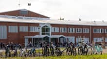 Koulun piha loppukesällä. Taustalle punaruskeatiilinen koulurakennus ja sen edessä koululaisia. Edustalla polkupyöriä telineessä.