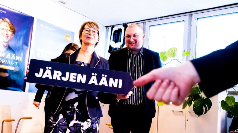 Sari Essayah järjen ääni vaalikyltti kädessään vaalivalvojaissa. Taustalla ehdokkaana ollut Kauko Pohjanen.