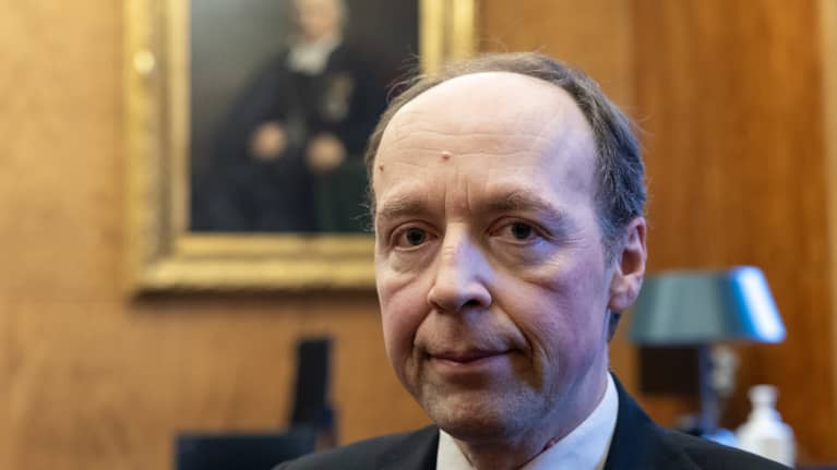 Vakavailmeinen mies katsoo kameraan työhuoneessa.