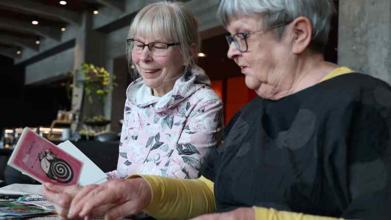 Tarja Seppänen ja Ritva Peltola istuvat pöydän ääressä ja katsovat postikortteja.