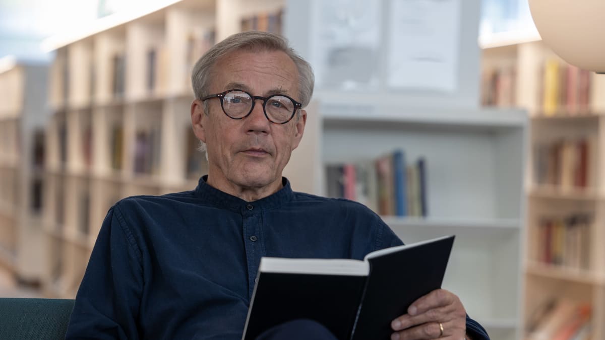 Harmaahiuksinen silmälasipäinen mies istuu kirja sylissään sinisellä sohvalla ja katsoo suoraan kameraan.