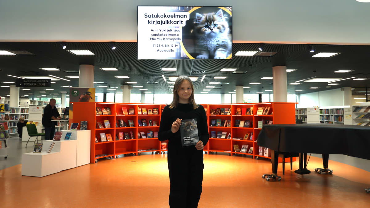 10-vuotias Armi Yoki esittelee kirjaansa osaksi oranssinsävyisessä kirjaston aulassa. Hänen yläpuolellaan näytöllä mainos kirjan julkistamistilaisuudesta.