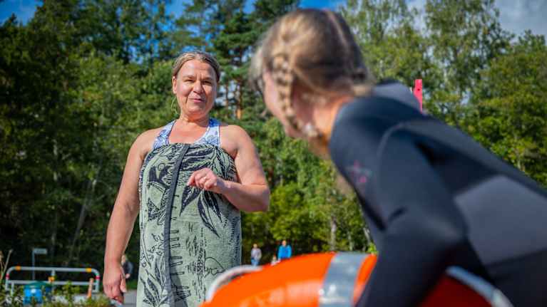 Kuvassa henkilö keskustelee rannalla toisen henkilön kanssa, joka on pukeutunut märkäpukuun ja pitämässä pelastusvälinettä. Taustalla näkyy vehreää metsää ja sininen taivas.