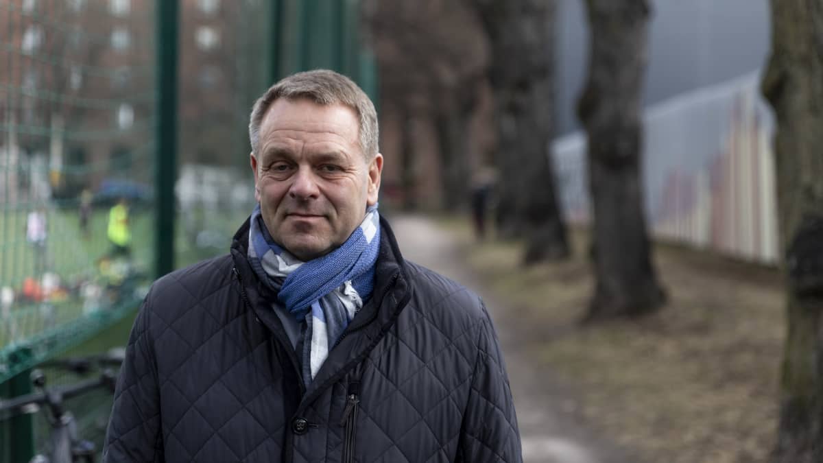 Jan Vapaavuori katsoo kameraan. Taustalla puilla reunustettu polku ja jalkapallokenttä. 