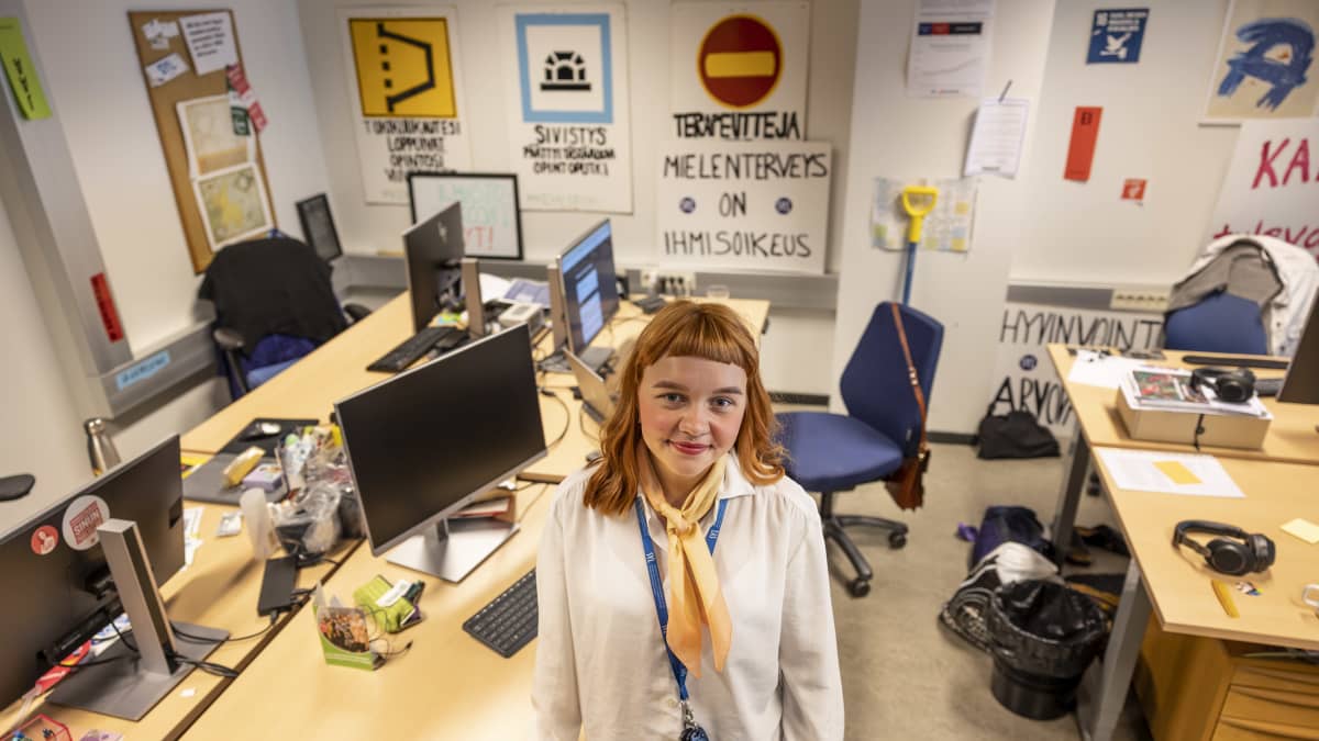 Jenni Suutari seisoo SYLin hallituksen toimistolla. Taustalla työpöytiä, tietokoneita ja julisteita, joista yhdessä lukee "Mielenterveys on ihmisoikeus".