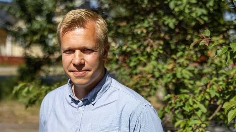 En man med ljusblå skjorta står utomhus framför grönskande träd.