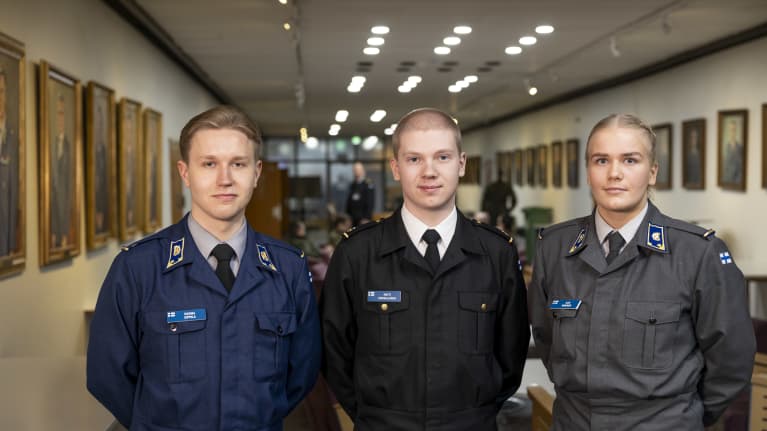 Kadetit Kuisma Sippola, Matti Koikkalainen ja Assi Nikkinen seisovat vierekkäin univormuissaan pitkällä käytävällä, jonka seinustoilla kultakehyksisiä tauluja.