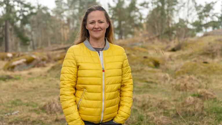 En person (Annika Kolster) med gul ytterjacka står utomhus i en skogsmiljö.