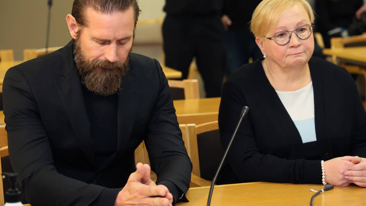 Mustiin pukeutunut, parraskas laulaja Lauri Tähkä ja asianajajansa Riitta Leppiniemi lähikuvassa istumassa oikeudessa päydän takana. Tähkän katse luotu alas, kädet yhdessä pöydällä.