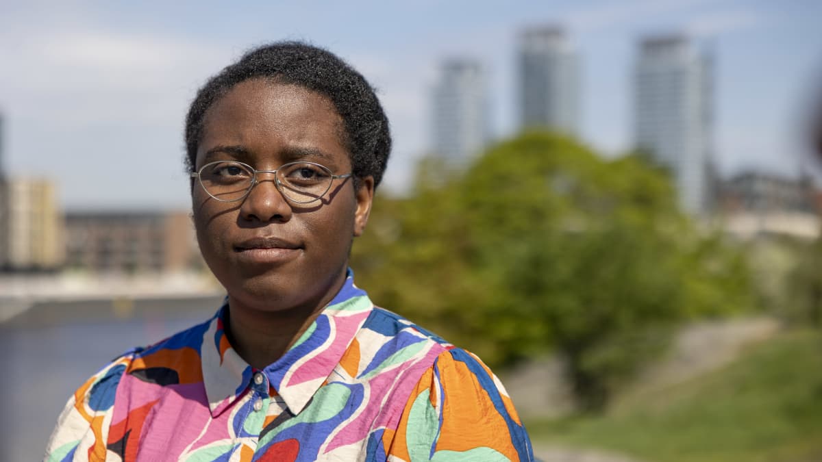 Jenny Kasongo katsoo kameran ohi, taustalla vihreä puu ja Kalasataman tornitalot.