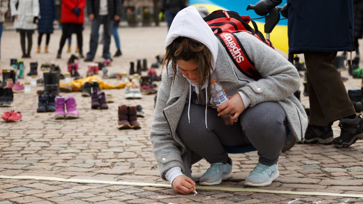Nainen kirjoittaa liidulla katukivetykseen. Taustalla näkyy maahan aseteltuja lasten kenkäpareja.