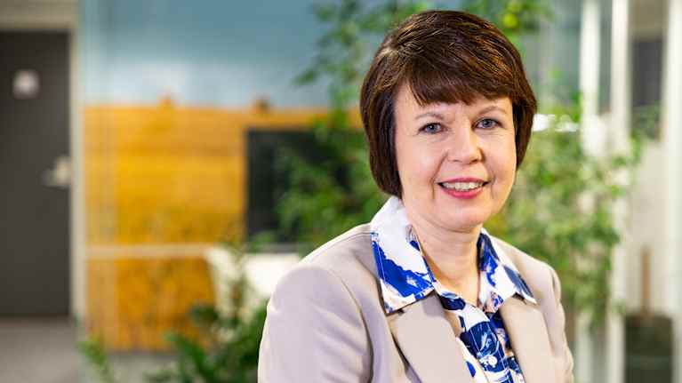 Sari Forsman-Hugg i en beige kavaj och blommig skjorta tittar in i kameran. I bakgrunden syns en växt och en tv.