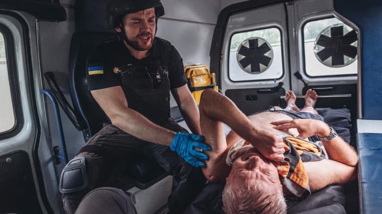 Ensihoitaja pitelee haavoittunutta siviiliä ambulanssin kyydissä. Ensihoitajalla on kumihanskat, luotiliivi ja kypärä. T-paidan hihassa on Ukrainan kelta-sininen lippu.
