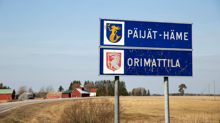 Orimattilan kaupunkikyltti ja Päijät-Hämeen maakuntakyltti päällekkäin maantien varrella. Taustalla paljas, keväinen maalaismiasema.