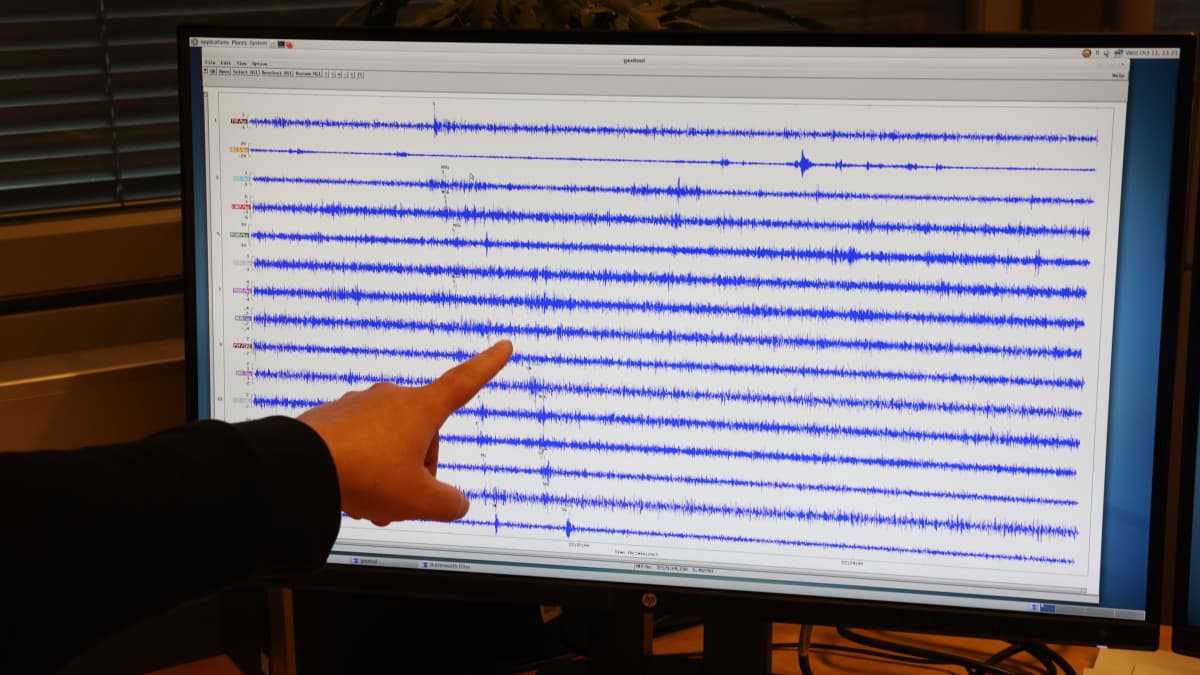 Seismologi osoittaa sormella sinisiä mittauskäyriä tietokoneen näytöllä. Taustalla kellertävä valo toimistohuoneessa.