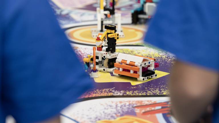 Legopalikoista tehty hahmorakennelma seisoo robotiikkakisan alustalla kahden siniseen t-paitaan pukeutuneen lapsen välistä.