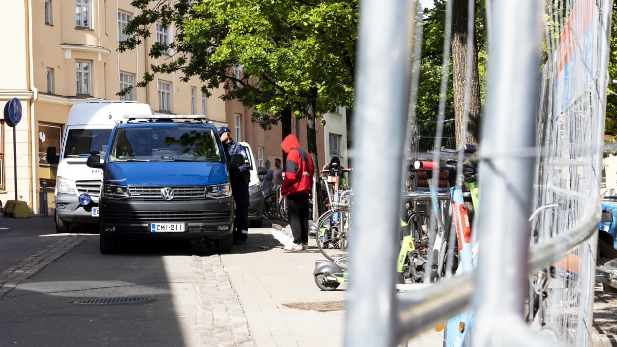 Poliisi kurkottaa jotain poliisiautosta. Huppupäinen siviili seisoo vieressä.