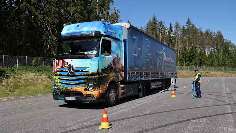 A man in uniform stands in a parking lot on a summer day, looking at a blue and orange truck with trees in the background.