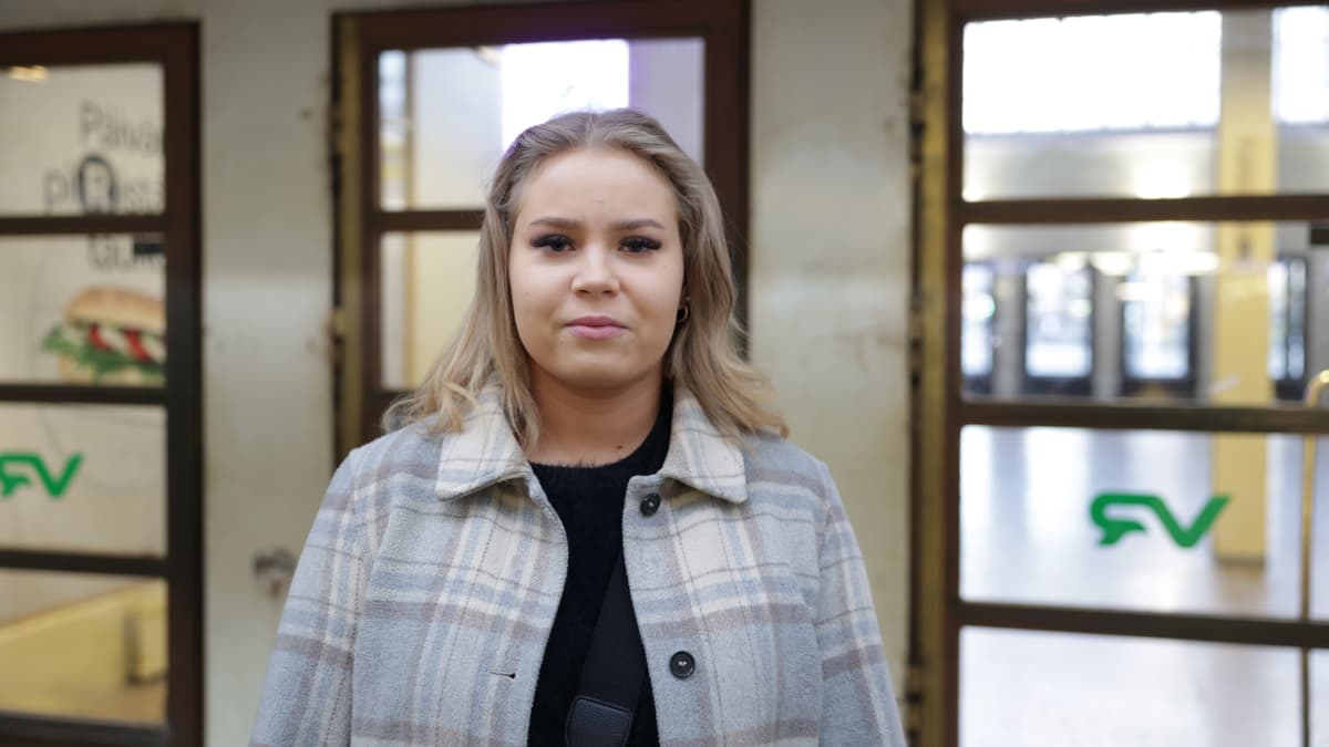 Elina Miettinen seisoo Tampereen rautatieasemalla, katsoo kameraan hymyillen, päällään vaalea takki. 
