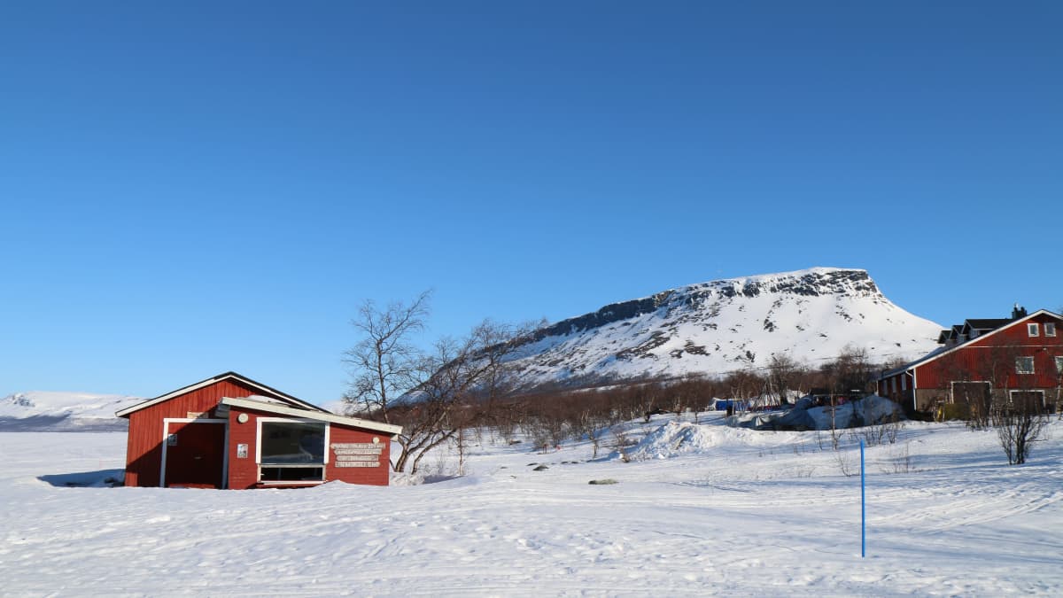 A red wooden cabin located on the beach of the Kilpijärvi Lake with Saanantunturi hill in the background.