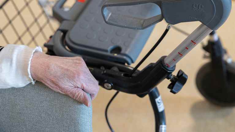 An elderly man&rsquo;s hand is gripping the armrest. A rollator is next to it.
