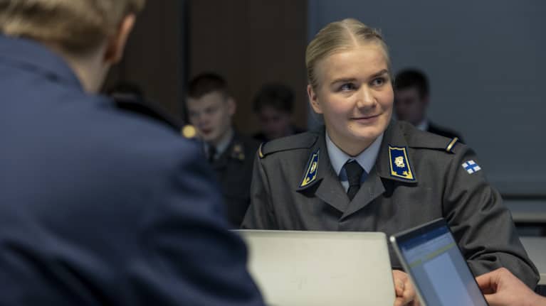 Harmaaseen univormuun pukeutunut kadetti Assi Nikkinen istuu läppärin ääressä opiskelutovereiden kanssa ja katsoo sivuun hymyillen.