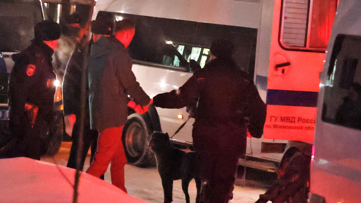 Himkin pöliisiaseman pihamaa. Kolme poliisi ohjaa Aleksei Navalnyia pakettiautoon. Auton takavalot värjäävät maiseman punaiseksi. Yhdellä poliisilla on koira hihnan päässä.