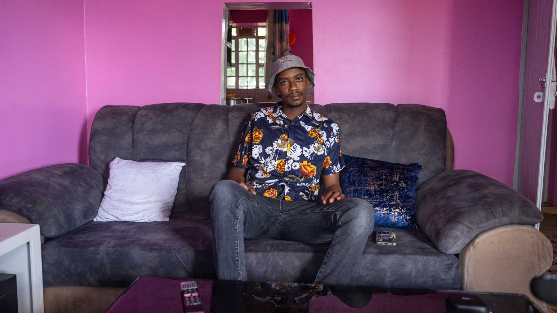 Voimakkaan vaaleanpunaiseksi maalatussa huoneessa iso harmaa sohva. Sen päällä istuu Tarab Ngugi kirjava paita ja kalastajahattu päällään.
