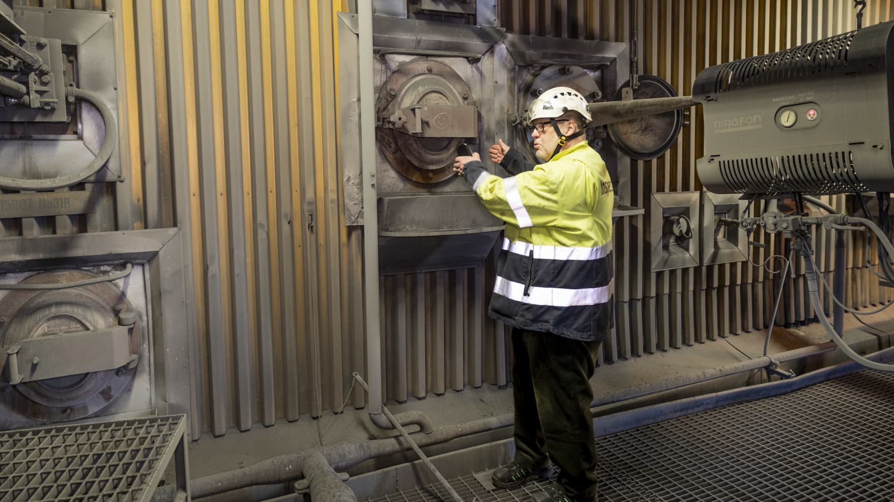 Työmaavaatteisiin pukeutunut mies osoittaa käsillä voimalaitoskattilan metalliseinässä olevaa ympyrän muotoista luukkua, jonka halkaisija on noin puoli metriä.