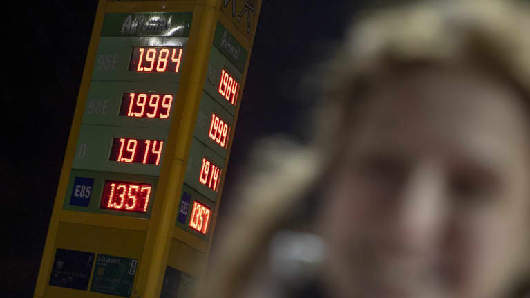 Minna Jaala seisoo huoltoaseman pihalla. Taustalla polttoaineiden hinnat. 