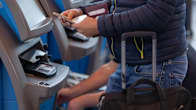 Close up photo of a man using an airport boarding pass machine, a suitcase is standing beside him.