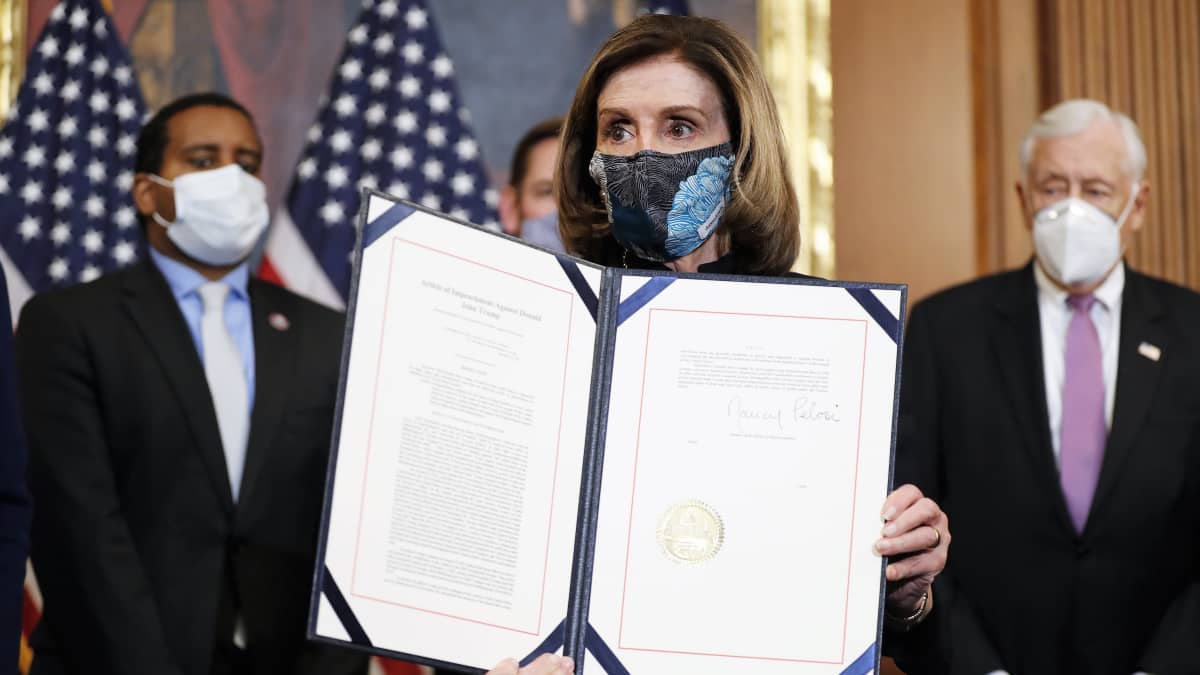Nancy Pelosi pitää käsissään asiakirjaa ja esittelee sitä kameroille. Hänellä on kasvoillaan maski. Taustalla näkyy kolme miestä tummissa puvuissa ja maskit kasvoilla. Huoneen seinän vierellä on Yhdysvaltain lippuja.