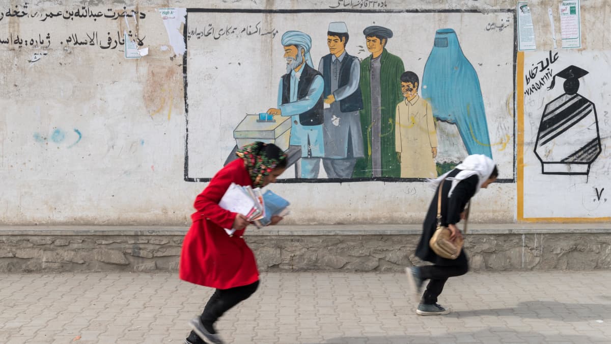 Kabulaistytöt matkasivat tuulessa kotiin koulusta joulukuussa 2017. Taustalla muuriin maalatussa mainoksessa kannustetaan miehiä ja naisia äänestämään.