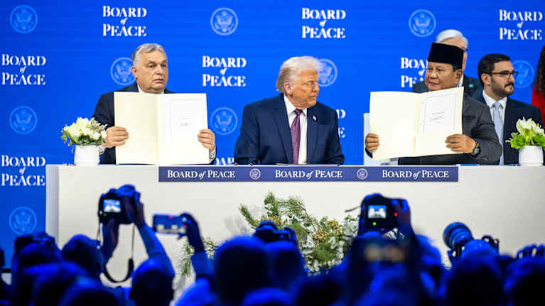 Three men sit at a table on a stage, two of them holding up documents and showing them to the audience seen in the foreground, some taking pictures..